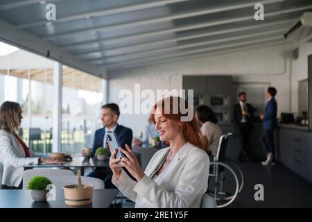 Woman using smartphone in break zone at work with colleagues eating. Concept: coworking, business, rest Stock Photo
