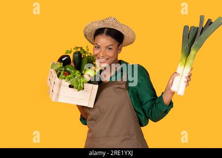 Black Farmer Lady Holding Box With Fresh Harvest, Yellow Background Stock Photo