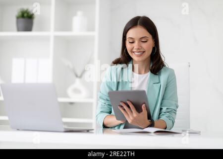 Cheerful millennial caucasian lady manager in suit typing on tablet at workplace with laptop, enjoy business Stock Photo