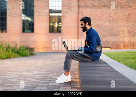 Indian Bearded Man Typing On Laptop Sitting Outdoor Stock Photo