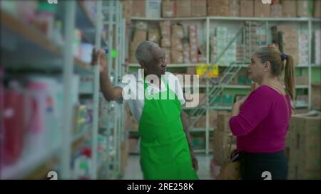 A male black senior employee listening to female customer leaning on shelf and wearing green apron, supermarket grocery store small business setting Stock Photo