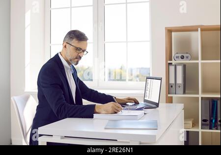Male accountant or bookkeeper working in office, using laptop, and taking notes in accounting book Stock Photo