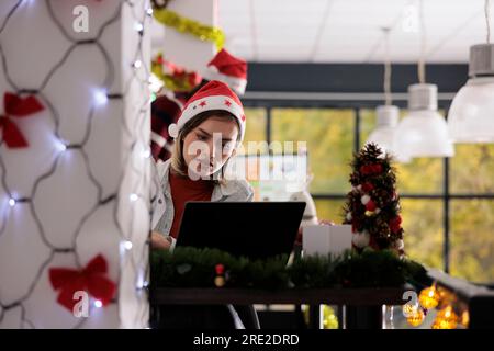 Focused businesswoman working on project tasks sitting at desk in Christmas themed workspace. Experienced employee researching data on laptop in festive decorated office during winter holiday season Stock Photo