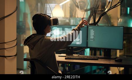 Asian hacker creating malware with vr glasses at night, using virtual reality to hack government server. Young criminal doing cyber espionage on dark web network, illegal phishing. Stock Photo