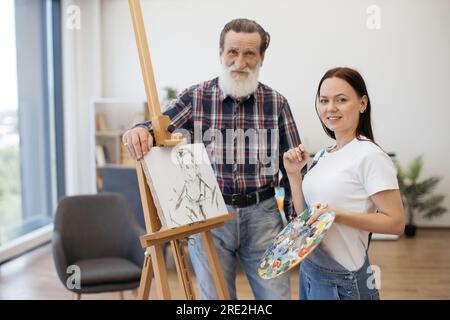 Cheerful senior gentleman and young female in casual outfits posing near easel with male portrait on canvas. Skilful amateur painter finishing head an Stock Photo