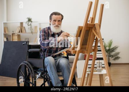 Portrait of happy senior man on retirement finding inspiration and drawing picture on canvas with watercolors. Charismatic aged man with disability looking straight in camera and smiling. Stock Photo