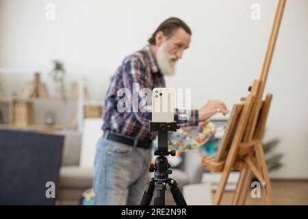 Casually dressed grey-bearded artist recording video using smartphone on tripod while painting portrait on easel. Concept of older generation using mo Stock Photo
