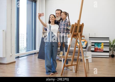 Happy family of three painters making selfie on modern cellphone while working on new drawing together at home studio. Loving people enjoying spending pastime with each other. Stock Photo
