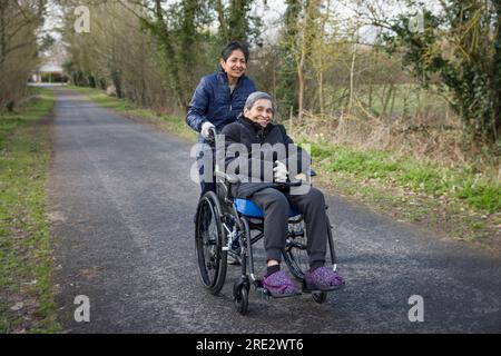 Asian Indian woman pushing her elderly mother in a wheelchair outdoors in winter, UK. May also depict a carer, care in the community Stock Photo