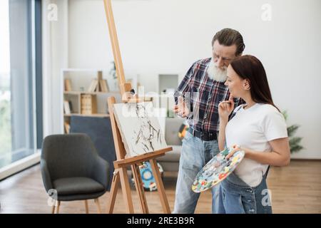 Focused senior man in casual outfit pointing at image sketch on fabric made by smiling woman in denim jumpsuit. Experienced art teacher analyzing elements of drawing during lesson in studio space. Stock Photo