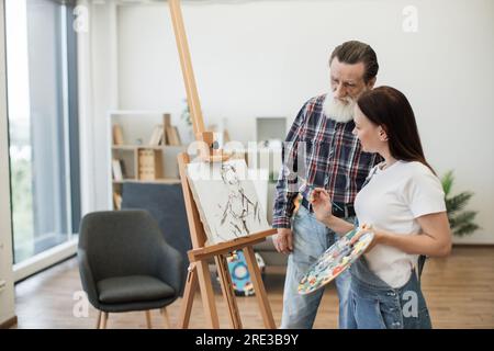 Focused senior man in casual outfit pointing at image sketch on fabric made by smiling woman in denim jumpsuit. Experienced art teacher analyzing elements of drawing during lesson in studio space. Stock Photo