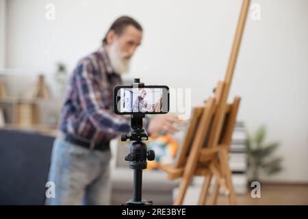 Blurred senior blogger broadcasting while painting new beautiful picture on easel at cozy art studio. Imposing elder male dressed in jeans and checkered shirt communicating with followers on internet. Stock Photo