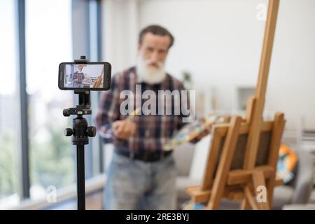 Blurred senior blogger broadcasting while painting new beautiful picture on easel at cozy art studio. Imposing elder male dressed in jeans and checkered shirt communicating with followers on internet. Stock Photo