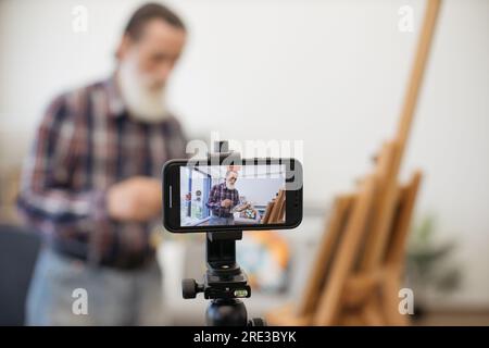 Blurred senior blogger broadcasting while painting new beautiful picture on easel at cozy art studio. Imposing elder male dressed in jeans and checkered shirt communicating with followers on internet. Stock Photo