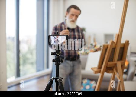 Blurred senior blogger broadcasting while painting new beautiful picture on easel at cozy art studio. Imposing elder male dressed in jeans and checkered shirt communicating with followers on internet. Stock Photo