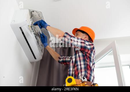 Worker installing or repairing air conditioner. Young man in uniform standing on ladder with toolbox and installing new air conditioner on wall. AC Stock Photo