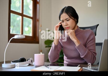 Stressful young Asian woman having a phone call with her coworker, discussing the project Stock Photo
