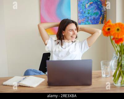 Happy satisfied latin woman rest at home office, sit with laptop hold hands behind head, dreamy young lady relax finished work, feel peace of mind look away , dream think of future success concept Stock Photo
