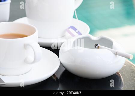 Cigarettes are placed in a white ceramic ashtray on a wooden table beside the coffee mug. Stock Photo