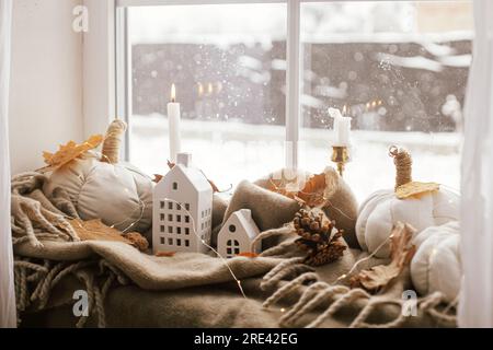 Cozy Autumn. Stylish pumpkin pillows, fall leaves, candle, lights and cute buildings decoration on brown scarf on windowsill. Autumn hygge, fall home Stock Photo