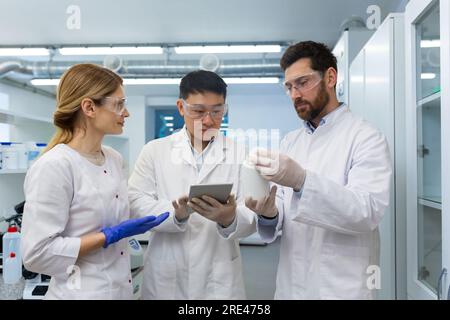 A team of three people, doctors, laboratory workers, scientists are standing inside a laboratory , researchers are communicating and discussing a scientific project, studying research material. Stock Photo