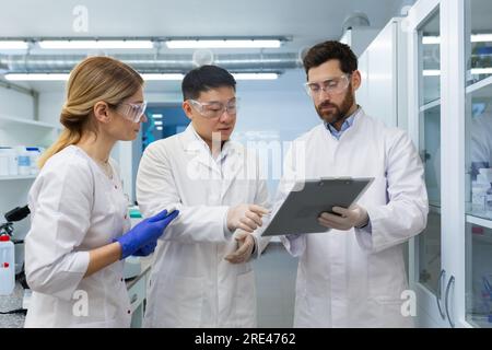 A team of three people, doctors, laboratory workers, scientists are standing inside a laboratory , researchers are communicating and discussing a scientific project, studying research material. Stock Photo