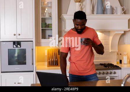 African american man drinking coffee and using laptop in white kitchen Stock Photo