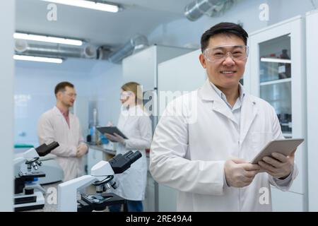 Asian scientist researcher in a white medical coat holds a tablet computer in his hands, the man works inside the laboratory. Stock Photo