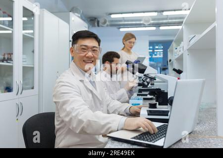 Portrait of asian lab technician, man with team group of people working in laboratory researching, man with microscope in white coat and goggles smiling and looking at camera. Stock Photo