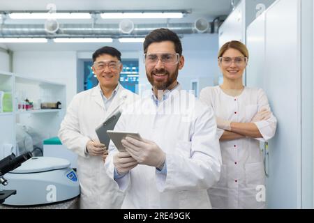 Successful team of scientists, three persons man and woman smiling and looking at the camera, portrait of a group of scientists in white medical coats inside a laboratory. Stock Photo