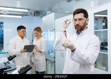 Mature experienced scientist inside laboratory researching and examining blood in test tube, man working with colleagues researchers in white medical coats. Stock Photo