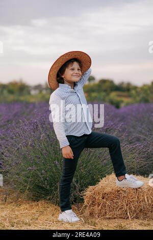 Joyful child boy in hat standing on hay bale against background of blooming lavender field and sky. Stock Photo
