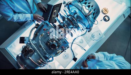 Two Professional Jet Engine Researchers Discussing Work, Using Laptop Computer and Checking Blueprints in Motor Manufacturing Facility. Scientists Stock Photo
