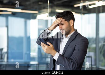 Upset hispanic businessman received online message notification with bad news, man frustrated thinking about important financial decision inside office at workplace. Stock Photo