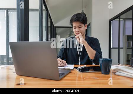 Business man asian using laptop and taking notes, writing business report, sitting at table at office Stock Photo