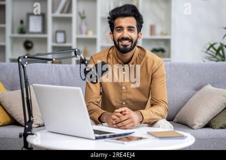 Portrait young Indian blogger influencer, man smiling and looking at camera, recording online podcast and video course from home, using professional microphone laptop, sitting on sofa in living room. Stock Photo