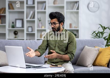 Serious focused and thoughtful Indian man talking on video call, man sitting at home in living room on sofa, using laptop for online communication with friends and colleagues. Stock Photo