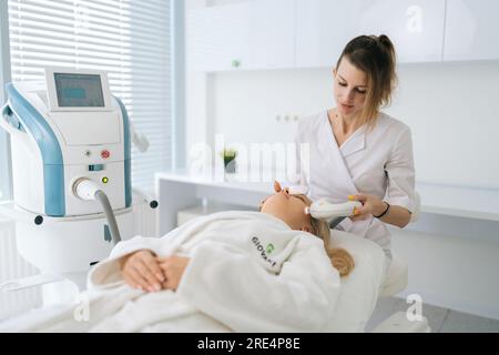 Portrait of female beautician doing hydro peeling procedure for woman in white bathrobe lying on medical couch in cosmetology clinic. Stock Photo