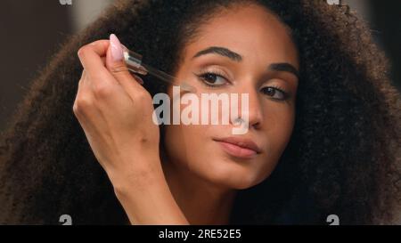 Close up female mirror reflection African American beautiful woman girl with perfect skin make-up apply oil face serum skincare product liquid Stock Photo