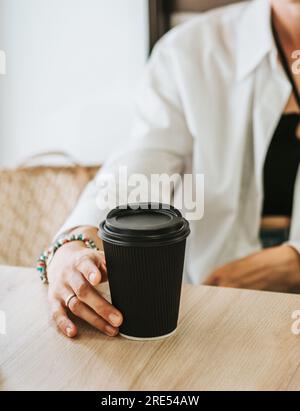 Black texture paper coffee cup in hand on table in cafe. Famale hand holds a disposable mug with a black lid. Advertising latte or tea. Stock Photo