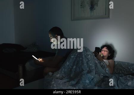 Dad and daughter are sitting on bed in teenage room, with smartphones in hand, using each of their smartphones separately, at night. Generation confli Stock Photo