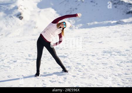 Slim woman in sportswear stretching arm sideway in snowy mountains Stock Photo