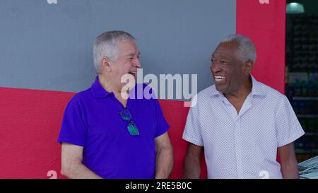 Joyful two diverse male senior friends smiling and laughing together leaning on sidewalk wall. A caucasian and African American older men interaction Stock Photo