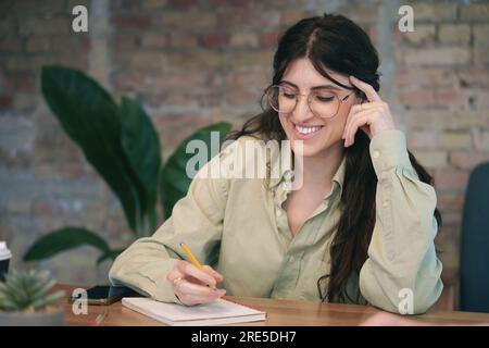 Smiling young brunette lady in glasses making notes in a notebook while working in the office. She working or studying. The startup, coworking Stock Photo