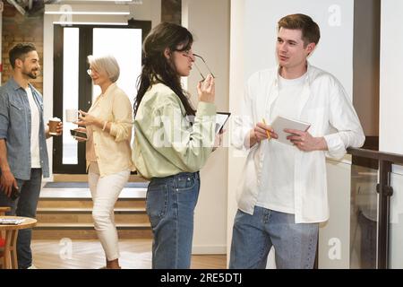 Young man and woman talking while standing in the office hall, two colleagues with coffee in the background. The startup, coworking, management Stock Photo