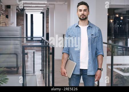 A male coworker posing at the camera while standing in the office and holding a laptop. The startup, coworking, and management concept Stock Photo
