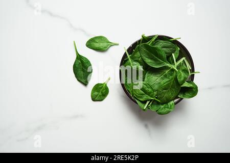 A bunch of Fresh baby spinach leaves in the in a black ceramic bowl on white marble background. Top view and flat lay photo with copy space Stock Photo