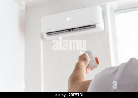 Hand holding remote control aimed at the air conditioner. Young man turning on air conditioner with remote control. Raising temperatures hot weather. Stock Photo