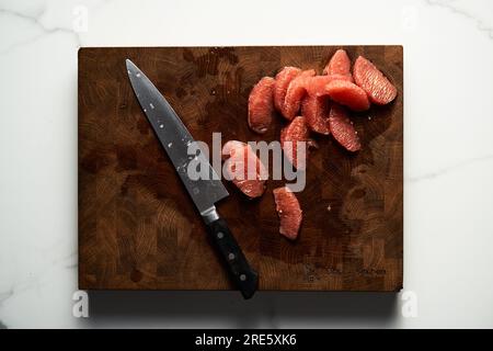 Peeled and sliced, divided in sections grapefruit on a wooden board with a knife next to it. The board rests on the marble countertop Stock Photo