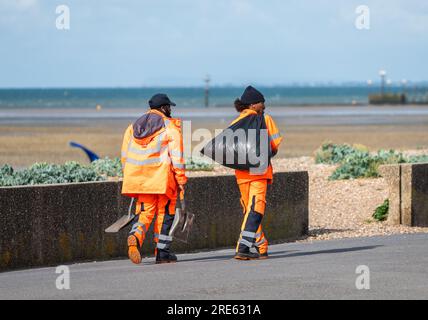 Pair of male workers, hi-vis clothing, walking along seafront road, collecting rubbish from litter bins & cleaning path on promenade in Summer, UK. Stock Photo